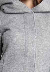 Grå hoodie med en mjuk textur, som har en stor huva, vertikala sömmar och en avslappnad passform. Materialet verkar lätt och bekvämt.