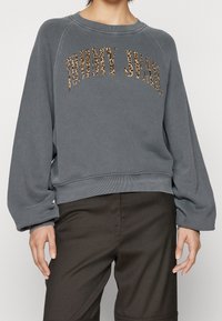 En person har på sig en grå oversized sweatshirt med "TOMMY JEANS" i leopardmönster över framsidan, tillsammans med mörka byxor.