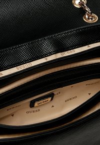 Borsa nera con texture e dettagli in catena, foderata in beige con il marchio "GUESS" e scompartimenti separati all'interno.