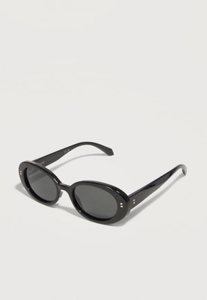 Μαύρα οβάλ γυαλιά ηλίου με σκοτεινούς φακούς και μικρές χρυσές τελείες κοντά στις αρθρώσεις, τοποθετημένα σε λευκή επιφάνεια.