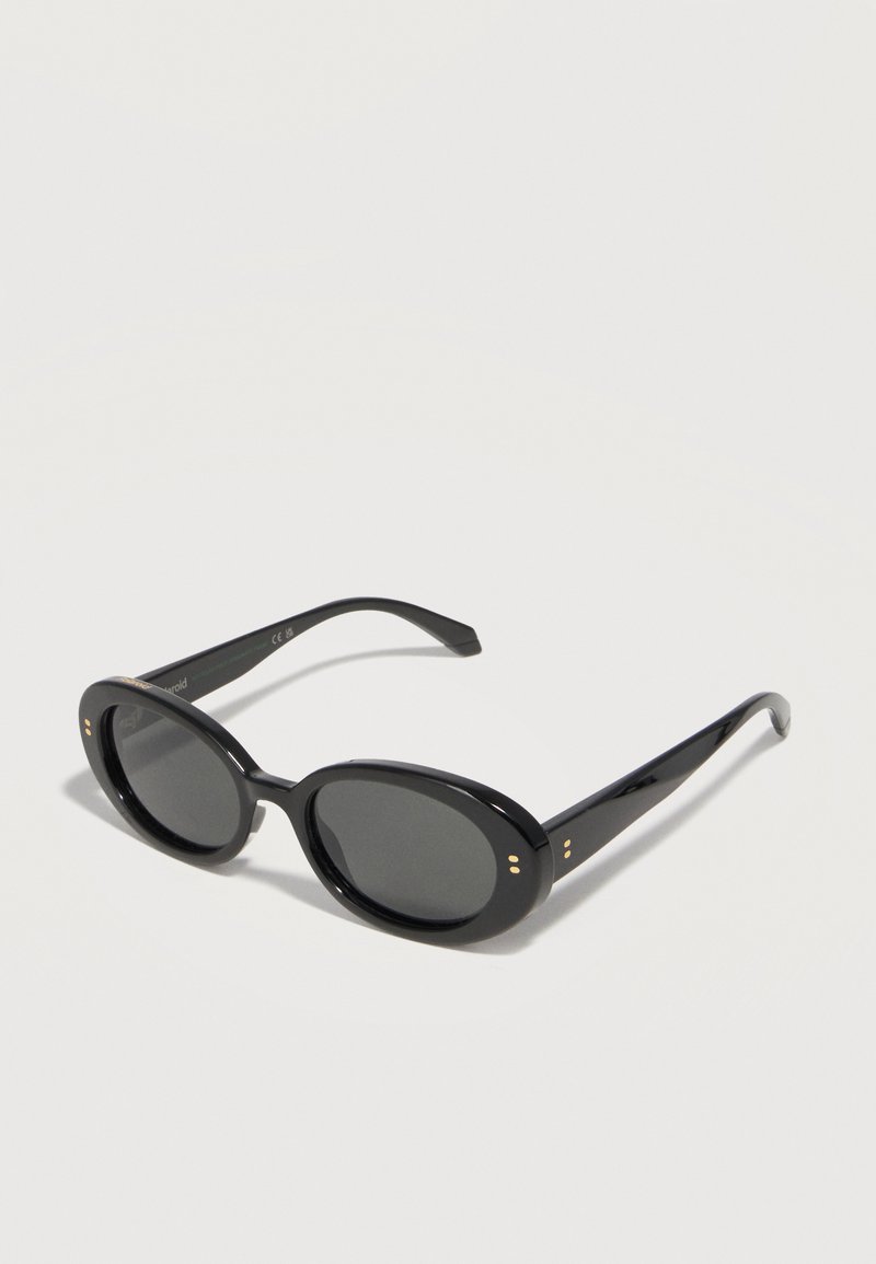 Μαύρα οβάλ γυαλιά ηλίου με σκοτεινούς φακούς και μικρές χρυσές τελείες κοντά στις αρθρώσεις, τοποθετημένα σε λευκή επιφάνεια.