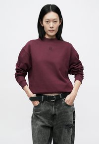 Mørkerød sweatshirt med ribbet halsringning og mansjetter, med en subtil "HUGO"-logo. Kombinert med høytlivde, slitte svarte jeans og et svart belte.