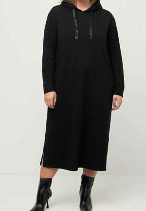 Femme portant une robe midi noire à manches longues avec capuche, fentes latérales et bottines noires à talons, debout sur un fond uni.