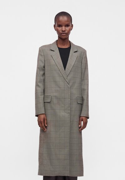 Femme portant un long manteau gris à carreaux par-dessus un haut noir, debout devant un fond clair uni, regardant droit devant avec une expression neutre.