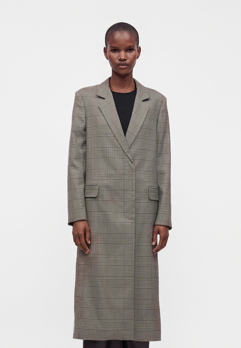 Femme portant un long manteau gris à carreaux par-dessus un haut noir, debout devant un fond clair uni, regardant droit devant avec une expression neutre.