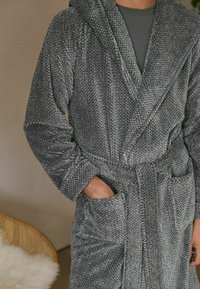 Grå strikbadekåbe med hætte, to forlommer og et bundet talje. Stoffet har et tekstureret mønster, som tilføjer dybde til designet.