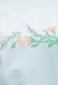 Υφάσμα ανοιχτό μπλε με μεγάλους λευκούς χαρακτήρες που είναι μερικώς ορατοί και παστέλ ροζ και πράσινες λουλουδένιες σχεδιαστικές διακοσμήσεις που εναρμονίζονται με το κείμενο.