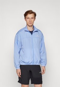 Mand iført en lysblå lynlåsjakke og sorte shorts, står mod en ensfarvet hvid baggrund, vender fremad med en neutral udtryk.