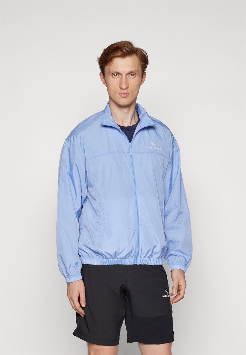 Mand iført en lysblå lynlåsjakke og sorte shorts, står mod en ensfarvet hvid baggrund, vender fremad med en neutral udtryk.