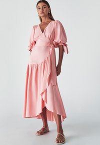 Long pink wrap dress with puff sleeves, v-neck, and asymmetric hem. Features gathered texture and waist ties. Worn with flat sandals.