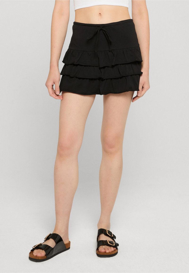 Femme portant une mini-jupe noire à volants avec taille à cordon et des sandales noires avec boucles dorées, debout sur un sol gris clair.