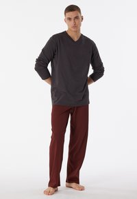 Langärmliges, anthrazitgraues V-Ausschnitt-Shirt, kombiniert mit rot gemusterten Schlafanzughosen. Das Model steht barfuß vor einem weißen Hintergrund.