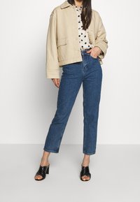 Giacca corta beige, blusa bianca con pois neri, jeans a vita alta blu e sandali neri con tacco decorati con borchie argentate.