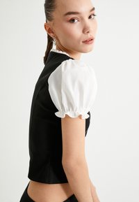Schwarzes, tailliertes Oberteil mit kurzen weißen Puffärmeln, Rüschensäumen und einem kurzen Design, das eine glatte Textur und klare Linien aufweist.