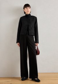 Schwarze strukturierte Jacke mit zwei Fronttaschen und runden Knöpfen, kombiniert mit schwarzen weit geschnittenen Hosen und schwarzen flachen Schuhen. Hält eine bordeauxfarbene Tasche.