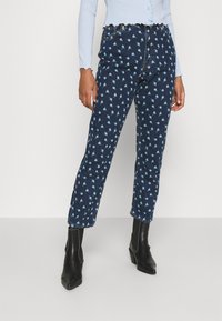 Personne portant un jean bleu foncé avec un imprimé floral bleu clair, un haut à manches longues en côtes bleu clair et des bottines noires à talons.