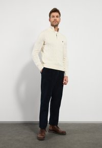 Cremfarbener Pullover mit Kabelstrickmuster und Reißverschlusskragen, kombiniert mit dunkelblauen Cordhosen und braunen Schnürstiefeln. Glatte Textur und taillierte Passform.