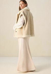 Femme portant un gilet en shearling crème par-dessus une blouse en dentelle blanche et une longue jupe fluide, se tenant contre un fond beige.