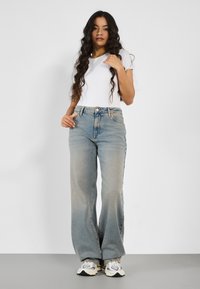 Nainen, jolla on pitkät kiharat hiukset, pukeutuneena valkoiseen t-paitaan, korkeavyötäröisiin leveälahkeisiin farkkuihin ja valkoisiin lenkkareihin, seisoo yksinkertaista taustaa vasten.