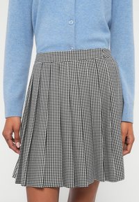 Fustă plisată alb-negru cu model houndstooth, realizată dintr-un material texturat, prezentată alături de un cardigan bleu deschis.