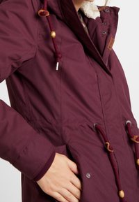 Frau trägt einen weinroten Wintermantel mit verstellbarem Kapuzenzug, Holzperlen, Druckknöpfen und einem mit Fleece gefütterten Kragen, Hand in der Tasche.