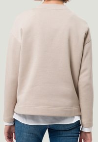 Beige, měkká mikina s kulatým lemem, dlouhými rukávy a žebrovanými manžetami. Viditelné švy na zadní straně; nošena přes bílou košili.