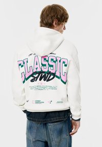 Witte hoodie met grote "CLASSIC" grafische afbeelding in roze en groen op de achterkant, met extra tekst en steraccenten. Draagt een spijkerbroek.