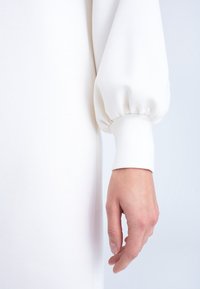 Vit klänning med puffärmar och en åtsittande manschett. Tyget ser slätt och mjukt ut, med en subtil glans. Hand placerad vid sidan.