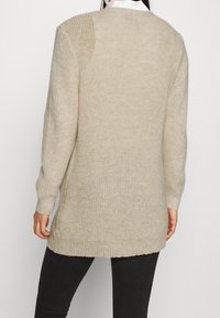 Knee-length beige knitted sweater with ribbed cuffs and hem, featuring a textured pattern and slightly oversized fit.