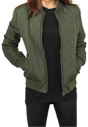 Vrouw die een opengeritste olijfgroene gewatteerde bomberjack draagt over een zwart shirt en zwarte broek, met handen in de zakken van het jasje.