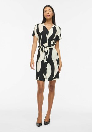 Femme portant une robe à manches courtes, à motifs noirs et blancs, boutonnée jusqu'au genou, avec une ceinture nouée à la taille et des talons hauts noirs.