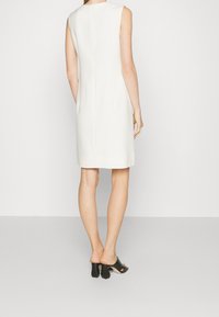 Robe blanche sans manches à coupe droite, avec une texture lisse et un design minimaliste. Portée avec des talons blocs noirs à bouts ouverts.