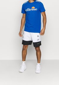 Camiseta azul con el logo de "ellesse" y pantalones cortos deportivos blancos y negros con el swoosh de Nike, combinados con zapatillas blancas y calcetines blancos.