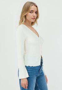 Cardigan bianco a costine con scollatura a V, maniche lunghe a campana e orlo scallopato. Caratterizzato da chiusura con bottoni sul davanti e texture morbida.