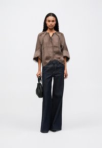 Frau mit langen, lockigen Haaren, die eine lässige braune Bluse und weite, dunkelblaue Jeans trägt, hält eine kleine schwarze Handtasche und steht vor einem weißen Hintergrund.