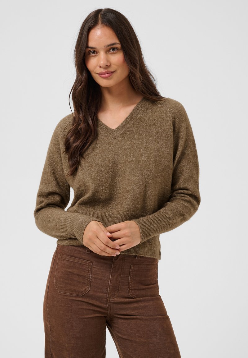 Jeune femme aux longs cheveux foncés portant un pull marron à col en V et un pantalon marron, debout devant un fond clair uni.