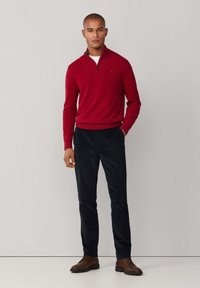 Roter Pullover mit Reißverschlusskragen, kombiniert mit dunkelblauen Hosen und braunen Lederschuhen. Das Ensemble zeichnet sich durch ein tailliertes Design und eine glatte Textur aus.