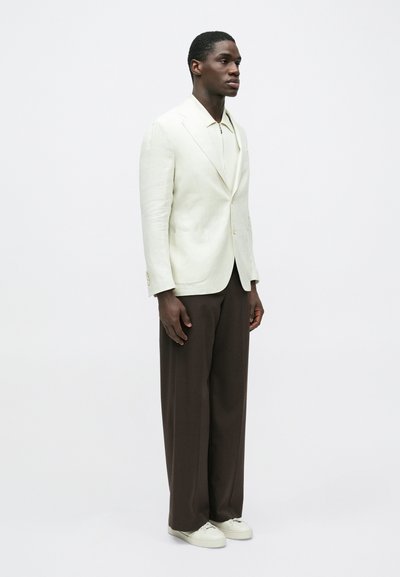 Homme debout portant un blazer blanc, une chemise zippée beige, un pantalon large marron et des baskets blanches sur un fond blanc uni.