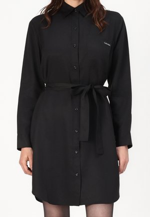 Femme portant une robe-chemise noire à manches longues avec col, ceinture nouée à la taille et petit logo blanc sur la poche poitrine.