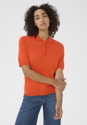 Donna con capelli ricci che indossa una polo arancione a maniche corte e jeans blu, che posa con un braccio incrociato, sfondo neutro.