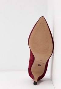 Chaussure à talon aiguilles en daim rouge à bout pointu, orientée vers le haut, montrant la semelle texturée couleur havane avec la taille 37 et la marque du fabricant.