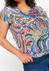 Donna che indossa un top corto con un motivo astratto multicolore e un collo a zip, insieme a jeans blu, con una mano appoggiata sui fianchi.