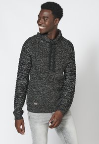 Felpa con cappuccio a maglia bianca e nera mélange, con collo alto, corde di regolazione e polsini a coste, abbinata a jeans grigi chiari.