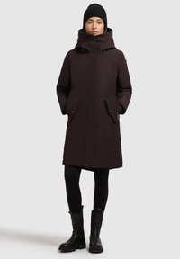 Parka aislante marrón oscuro con cuello alto, dos bolsillos delanteros y cierre de cremallera. Lleva puesto sobre unas mallas negras y botas de tobillo.