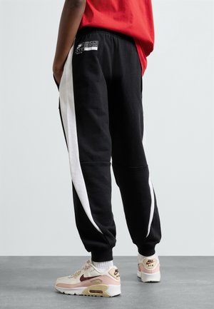 Pantalones de chándal negros con franjas laterales blancas, que presentan un corte holgado y una cintura elástica, combinados con zapatillas beige y burdeos.