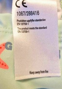 Vävt tygmärke med skötselanvisningar, vit bakgrund och svart text. Innehåller CE-märke och säkerhetsmeddelande: "Håll borta från eld."