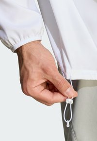 Hvid, letvægtsjakke med elastiske manchetter; håndjusterer en snor i kanten for pasform. Glat tekstur uden synlige mønstre.