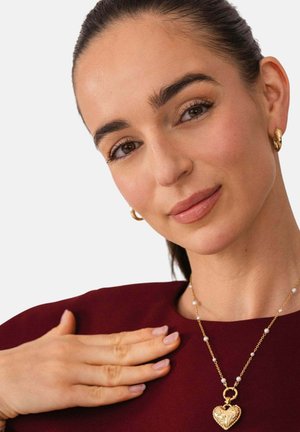 Frau mit dunklem Haar und goldenen Creolen, trägt ein burgundfarbenes Oberteil und eine goldene Halskette mit herzförmigem Anhänger, Hand auf ihrer Brust ruhend.