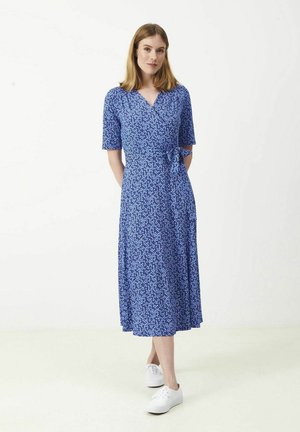 Kvinde iført en blå blomstret slå-om-kjole og hvide sneakers, stående med hænderne bag ryggen mod en ensartet lys baggrund.