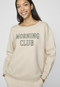 Sudadera beige con un escote amplio y mangas largas. Presenta "MORNING CLUB" en letras negras negritas en la parte frontal. Textura suave.